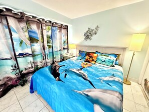 Une chambre Ă coucher avec un lit ornĂ© dâun motif reprĂ©sentant une plage tropicale, des rideaux illustrant une scĂšne de plage et deux lampes de chevet.