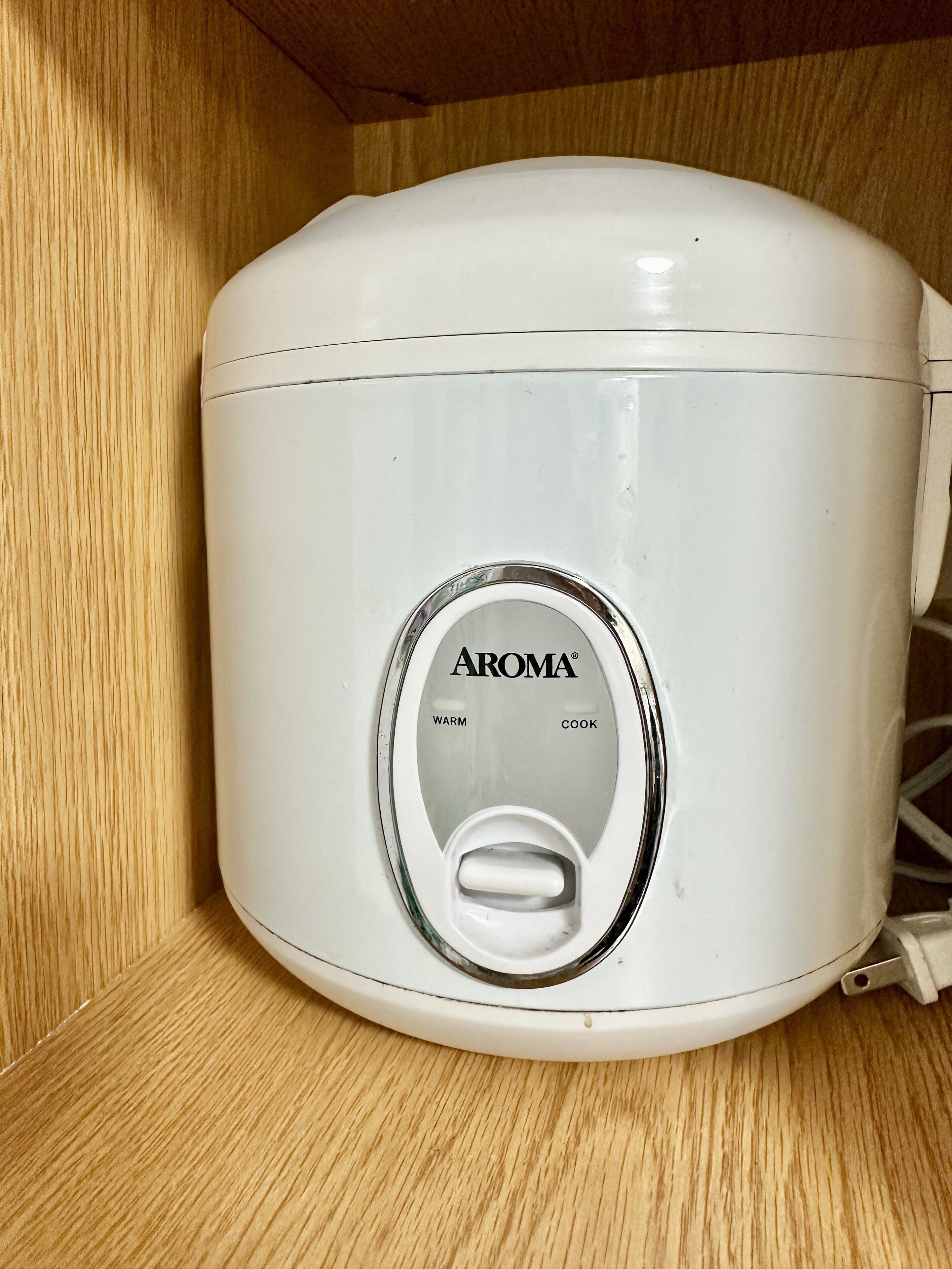 A white Aroma slow cooker with a control panel featuring 'WARM' and 'COOK' settings, placed on a wooden shelf.