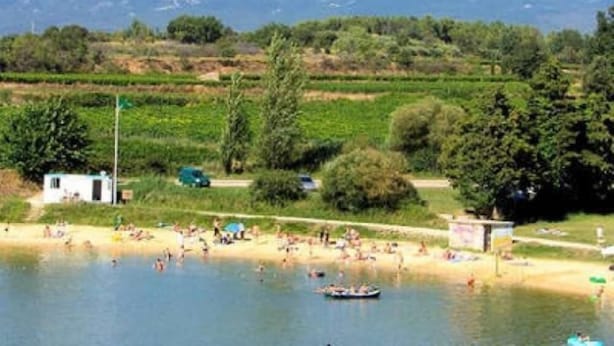 A lakeside view with a mountain in the background, a sandy beach, and people swimming and relaxing.