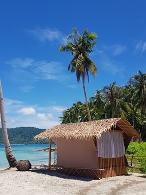 A beach hut with a thatched roof, situated on a sandy beach with palm trees and a clear blue sky in the background. - Backpackers Island Beach Camp - Adults Only (El Nido)