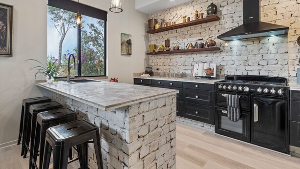 A kitchen with a brick wall, black cabinetry, and a large stove. There are stools and a window with a view of trees. - Jardin Tiny House (Beechmont)