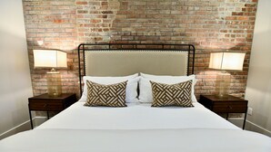 Una cama con edredón blanco y dos almohadones decorativos, flanqueada por mesitas de noche con lámparas, frente a un fondo de pared de ladrillo.