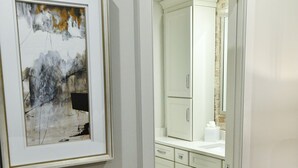 Un pasillo con una obra de arte enmarcada en la pared, suelo de madera y vistas a un baño con espejo y armarios.