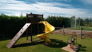 A playground with a wooden slide and climbing structure, surrounded by a fenced area and greenery. - LE PESCINE - Casa Oliveta: Spighe [OP] (Vada)