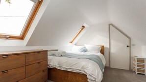 Ein Schlafzimmer mit schrägen Wänden, einem hölzernen Schminktisch, einem Bett mit weißer Bettwäsche und einem Oberlicht.