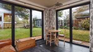 Ein modernes Zimmer mit großen Schiebeglasfenstern, die auf einen Garten führen. Es gibt eine hölzerne Bank, einen Tisch mit Stühlen und einen Deckenventilator.