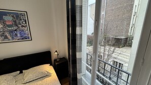 A bedroom with a bed, a nightstand, a lamp, a window with a view of buildings, and a framed picture on the wall. - Gorgeous Paris EIFFEL TOWER (Paris)