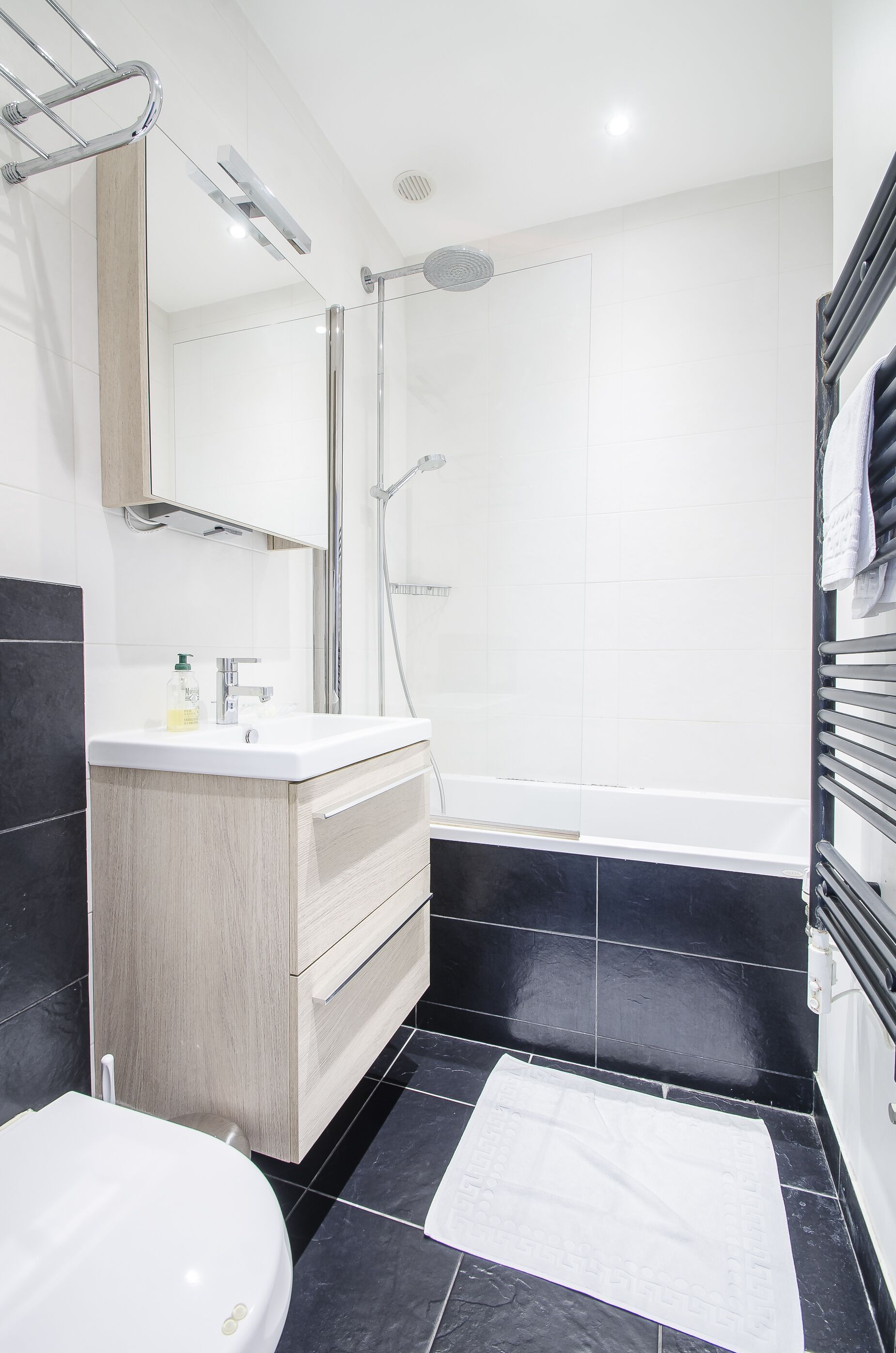 A modern bathroom with a white sink, wooden vanity, black tiled floor, and a white towel on the floor.
