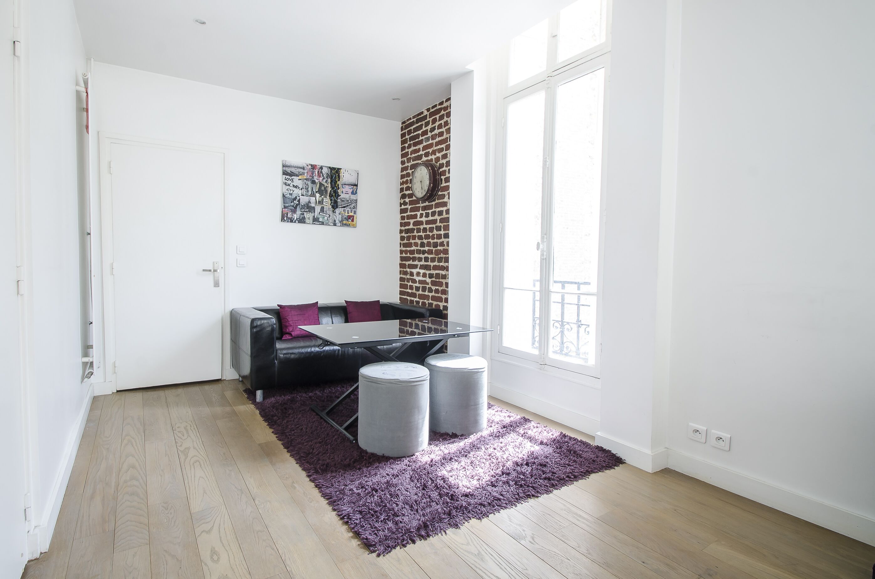 A modern living room with a black leather sofa, a glass coffee table, and two grey ottomans on a purple rug. A brick wall and a large window provide natural light.