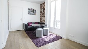 A modern living room with a black leather sofa, a glass coffee table, and two grey ottomans on a purple rug. A brick wall and a large window provide natural light. - Gorgeous Paris EIFFEL TOWER (Paris)