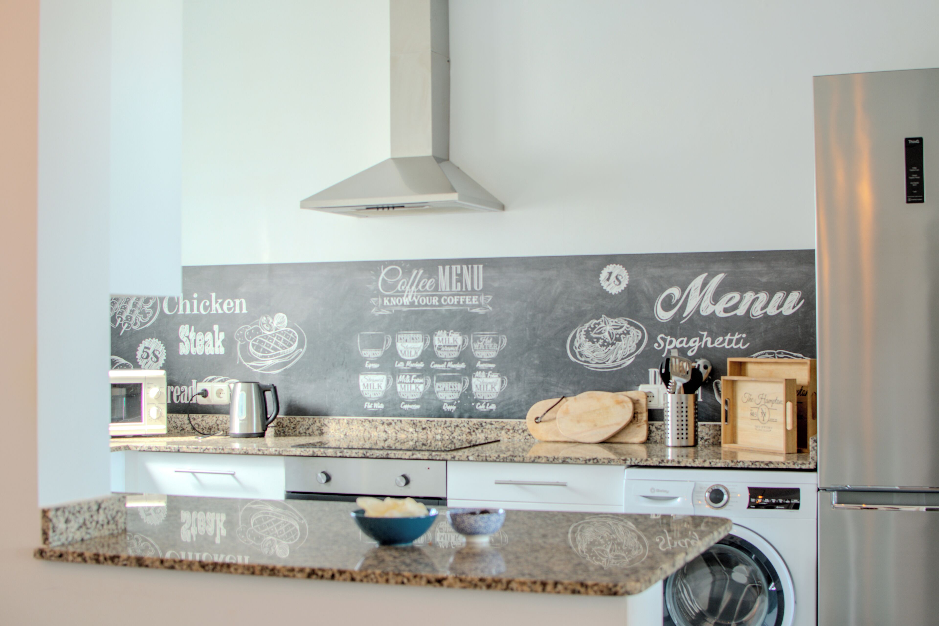 Una cocina con un menú en pizarra, microondas, cafetera y lavadora.