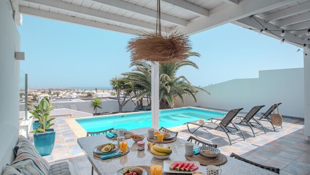 Ein Essbereich am Pool mit einem für das Frühstück gedeckten Tisch, mit Blick auf einen Swimmingpool und Palmen.