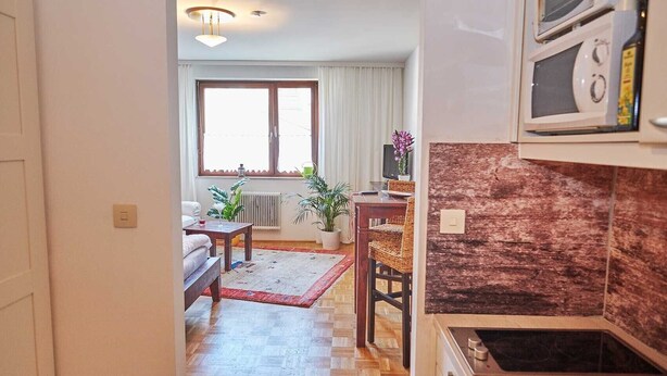 Un espacio habitable compacto con kitchenette, una zona de comedor con mesa y sillas, un sofá y un ventanal con cortinas.