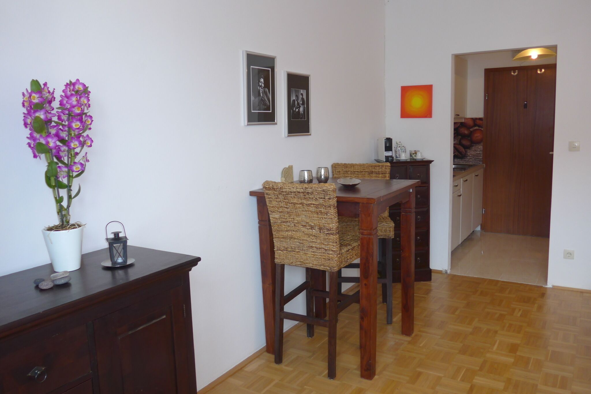 Ein Essbereich mit Holztisch und Korbstühlen, einer Topfpflanze und einem an der Wand hängenden Bilderrahmen.