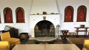 Een kamer met een open haard, gele banken, een tafel en rode, boogvormige doorgangen.