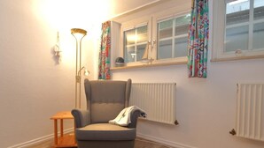 A room with a grey armchair, a small wooden table, a floor lamp, and a window with patterned curtains. - South beach 11 (Westerland)