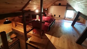 A loft bedroom with a sloped ceiling, wooden beams, a bed with a red blanket, a chair, a desk, and a window. - Welcome to Hautes-Pyrénées (Campan)