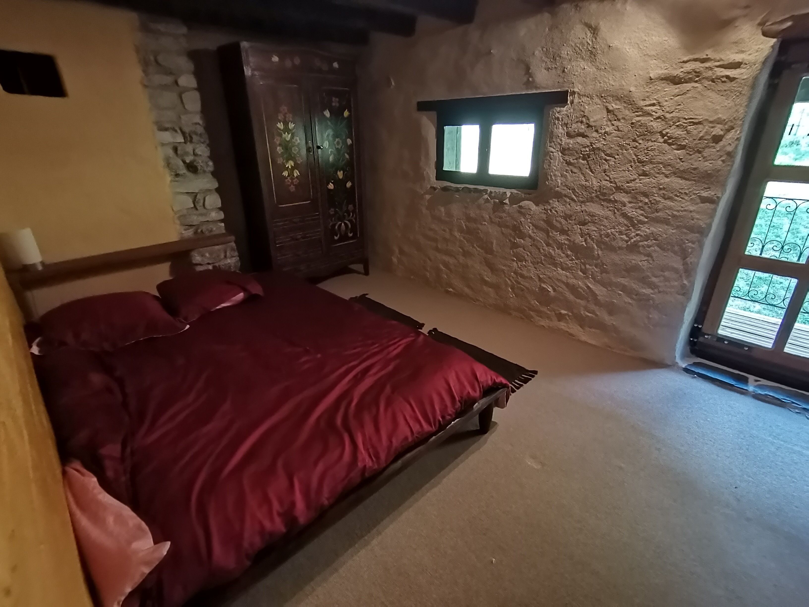A bedroom with a stone wall, a wooden bed with red bedding, a wooden wardrobe, and a window with a view.