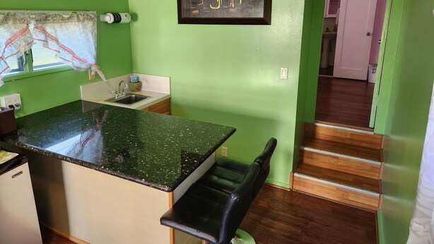 Una cocina con encimera verde, un lavamanos y una silla. En la pared, hay un cuadro con botellas de vino.