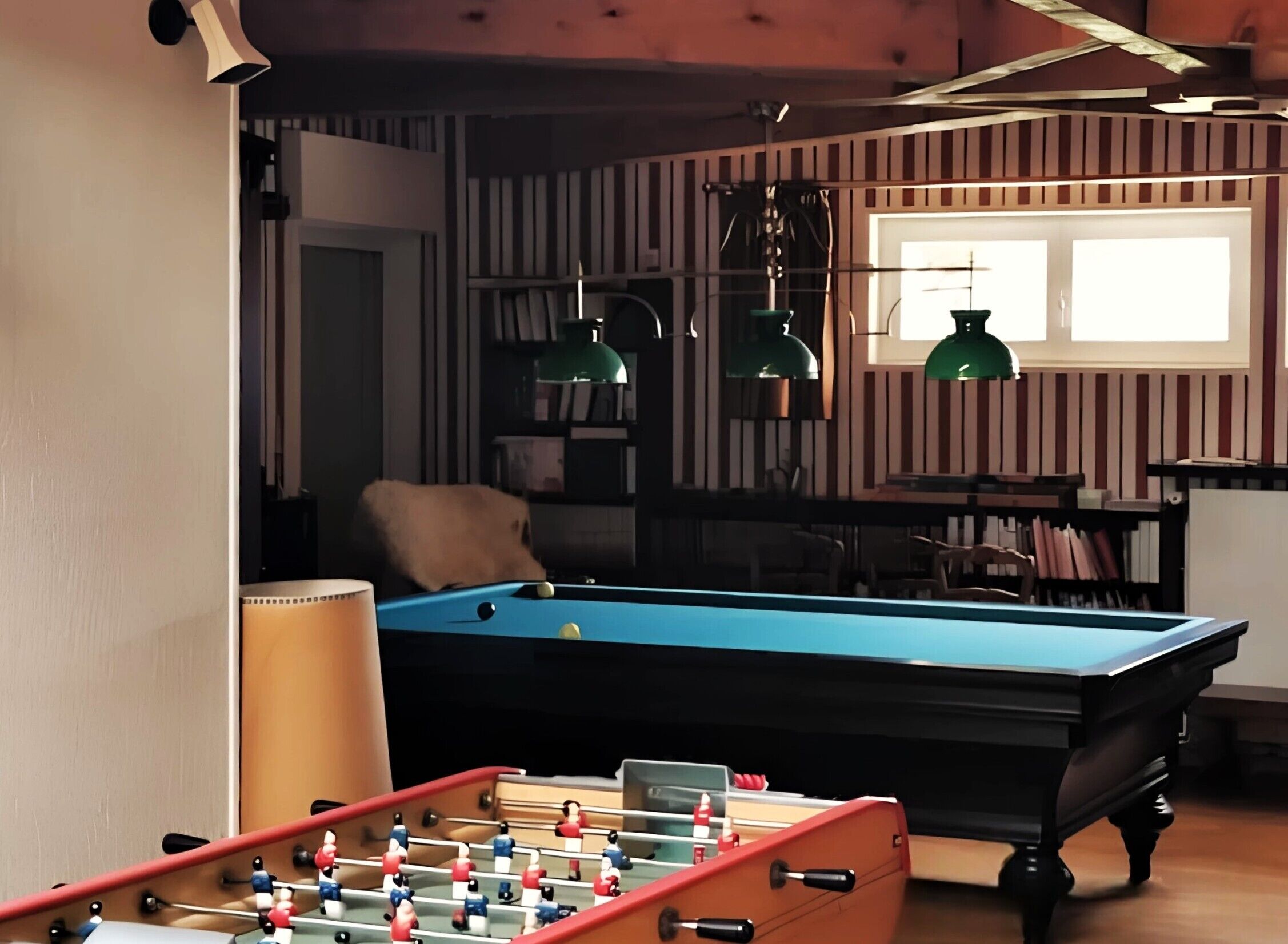 A pool table, a foosball table, and a window with striped curtains in a room with wooden beams and green pendant lights.