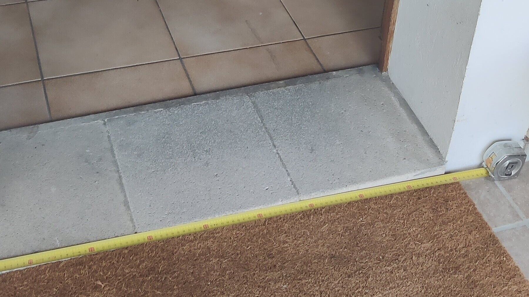 A corner of a room with a grey tiled floor, a measuring tape, and a corner with a white wall and a grey baseboard.