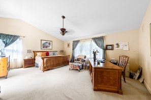 Ein geräumiges Zimmer mit einem großen Bett, einem Schreibtisch und einem Sessel. Es gibt einen Deckenventilator und ein Fenster mit Vorhängen.