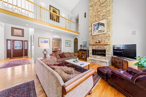 A spacious living room with a high ceiling, a stone fireplace, a large TV, and a comfortable seating area with a leather couch and patterned rugs.