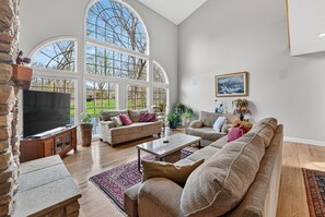 A spacious living room with a large TV, comfortable seating, and a view through large windows.