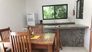 Una cucina con tavolo da pranzo in legno, sedie, frigorifero, una finestra vista alberi e un estintore al muro.
