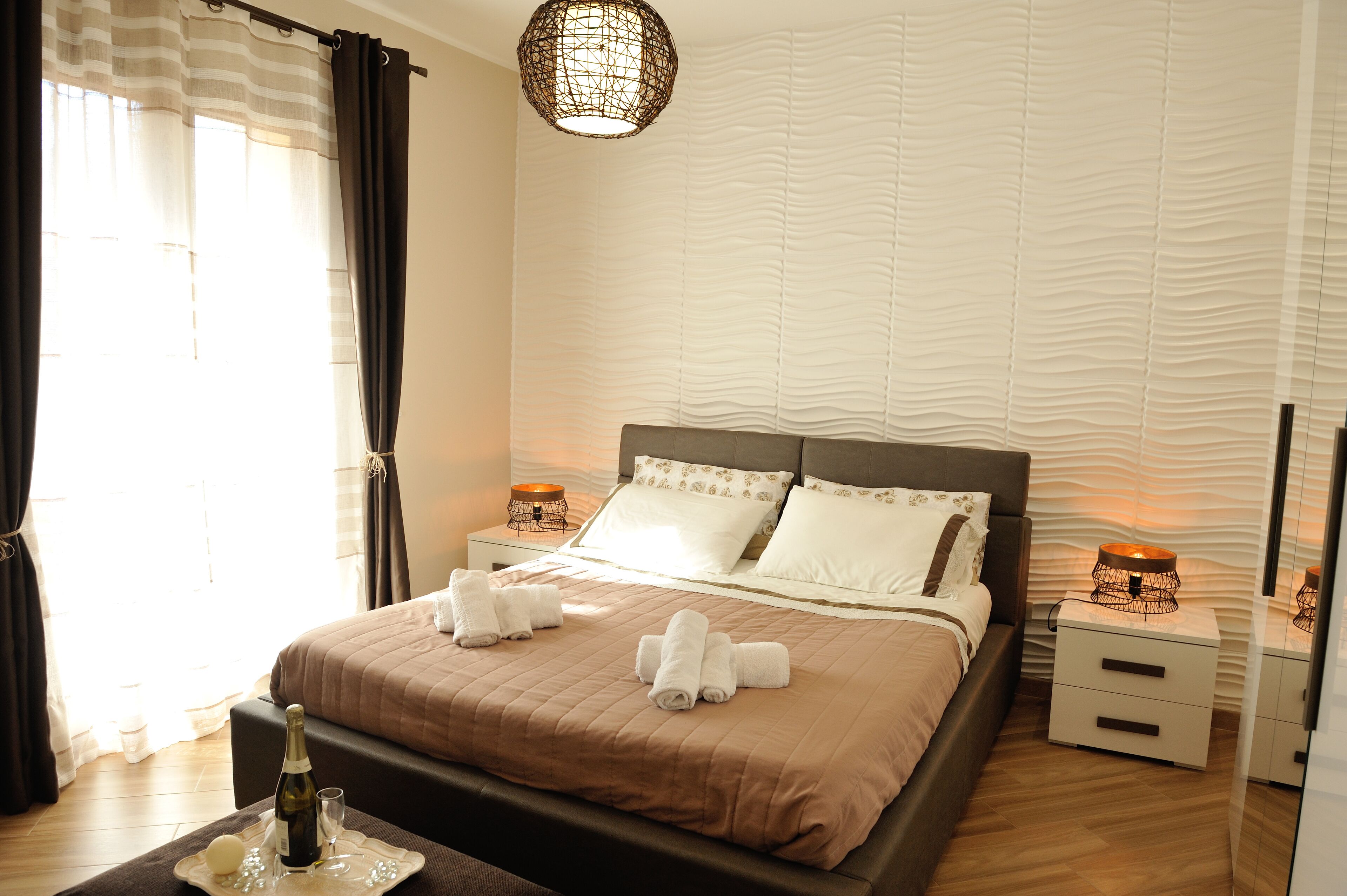 A neatly made bed with a brown bedspread, white pillows, and two white towels folded on the bed. There is a bedside table with a lamp and a bottle of champagne. The room has a modern design with a textured wall and a hanging light fixture.
