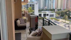 A balcony with a sofa and a small table, overlooking a cityscape with high-rise buildings and palm trees.