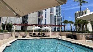 A swimming pool with lounge chairs and umbrellas, surrounded by high-rise buildings and palm trees.