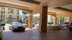 A covered parking area with a BMW parked in the foreground, a fountain, and a building with large windows in the background.