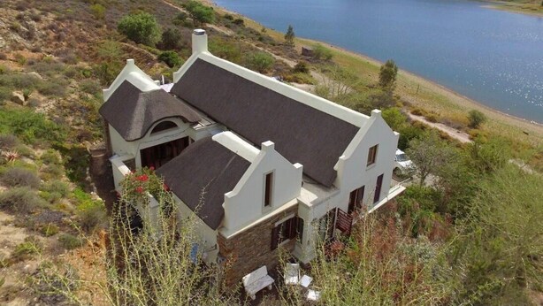A house with a stone foundation and a dark roof, surrounded by a natural landscape with a lake and hills in the background. - Xairu at Le Domaine Eco-Reserve (Country living) (Montagu)