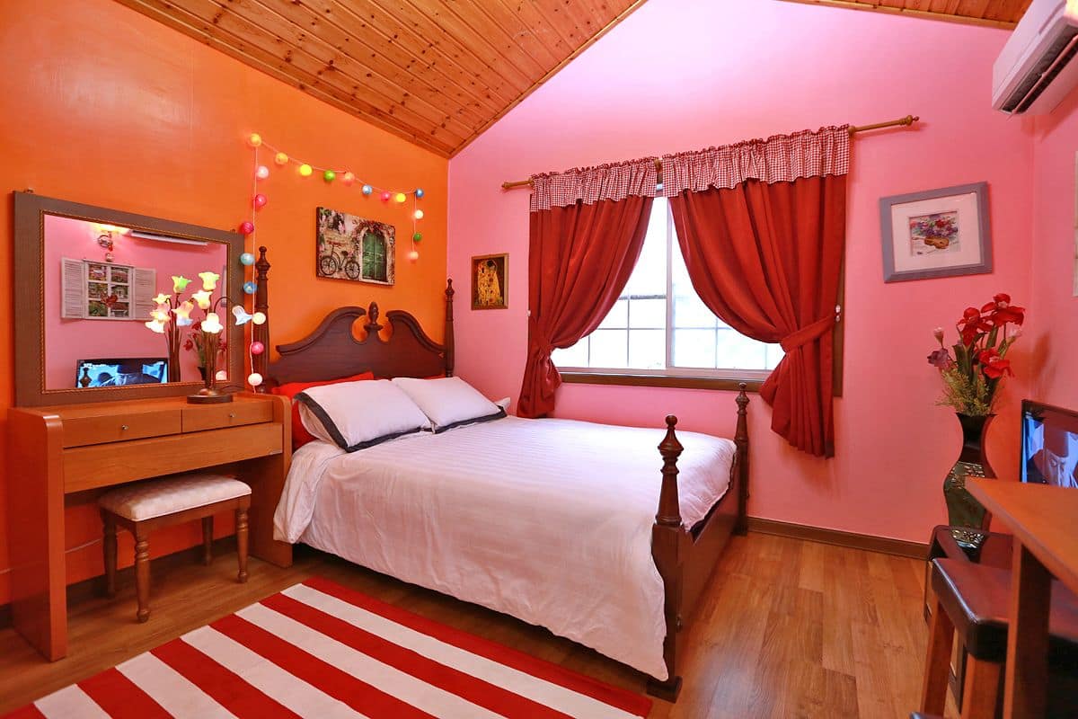 A bedroom with a wooden bed, a desk, and a chair. There is a window with red curtains and a mirror above the desk.