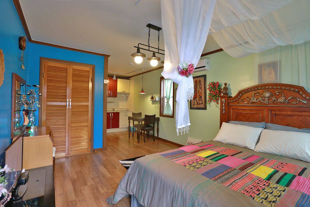 A bedroom with a large bed, a wooden headboard, and a canopy. There is a desk with a chair and a small table with a lamp. The room has wooden floors and a blue wall.