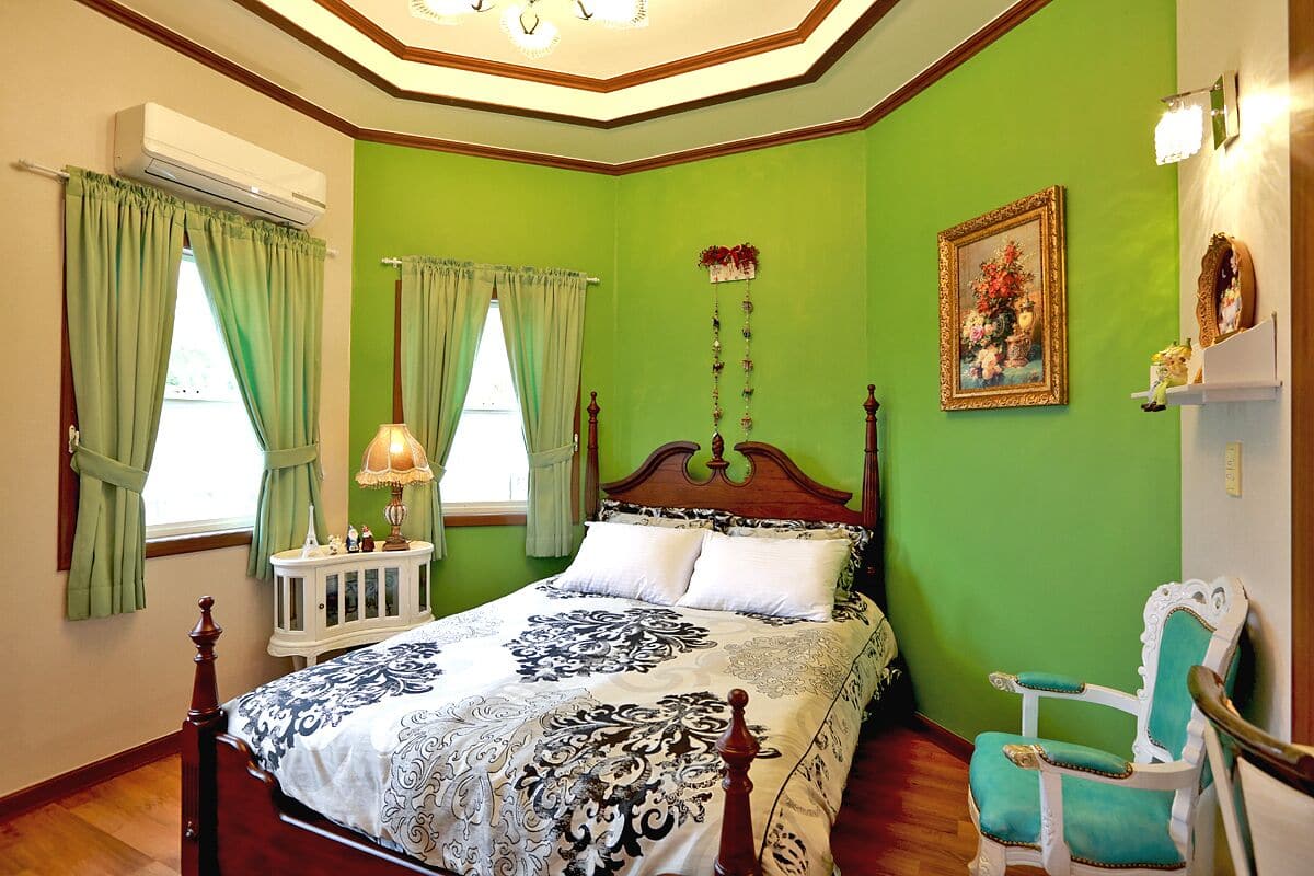 A bedroom with a green wall, a wooden bed, a white nightstand, a teal chair, and a painting on the wall.
