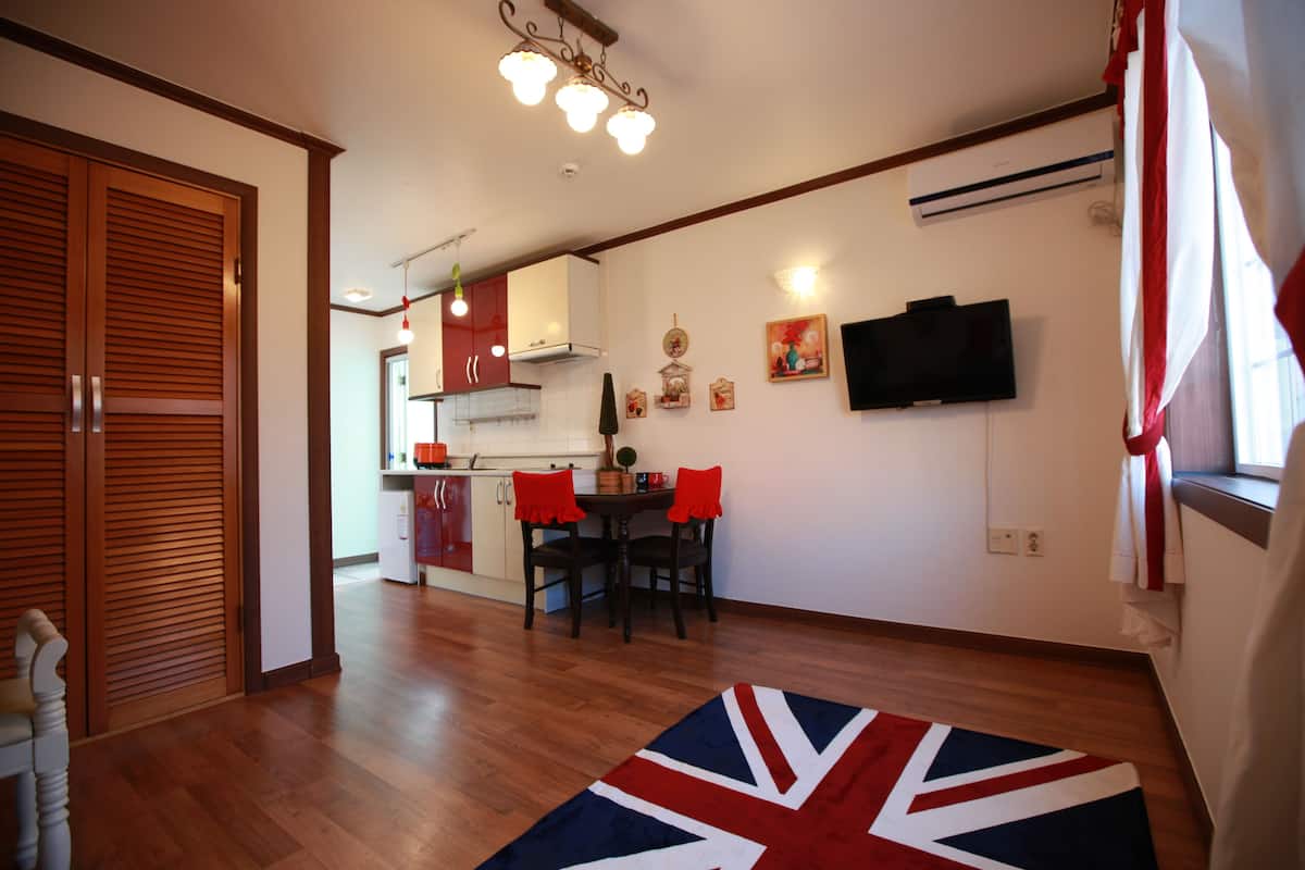 A modern kitchen with wooden cabinets, a dining area with red chairs, a Union Jack rug, and a flat-screen TV on the wall.