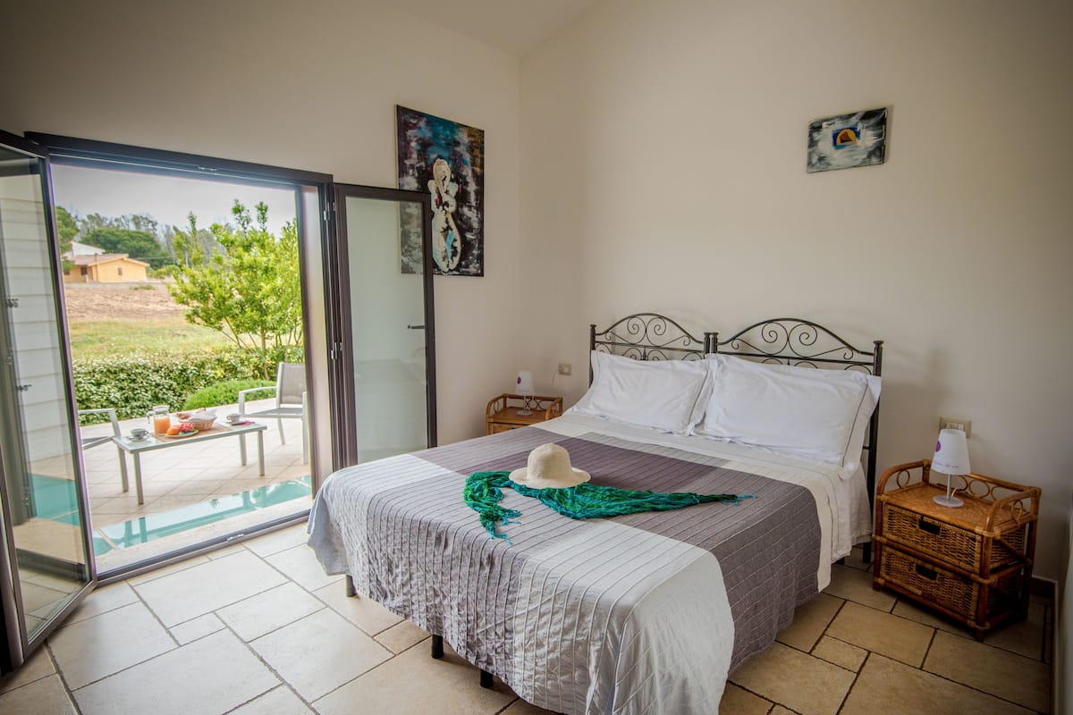 A bedroom with a bed, a nightstand, a wicker side table, a glass door leading to a patio, and a painting on the wall.