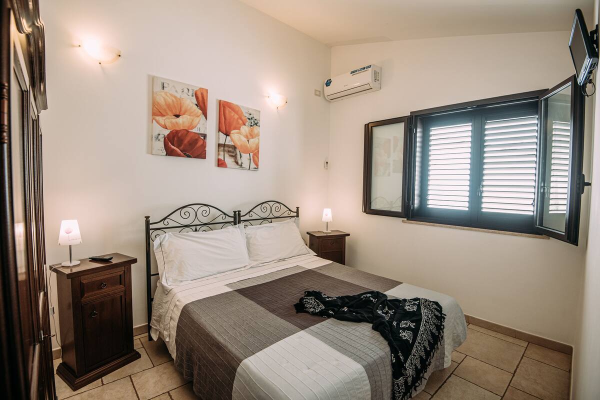 A bedroom with a bed, two bedside tables, a nightstand, a window with blinds, and wall-mounted air conditioning.