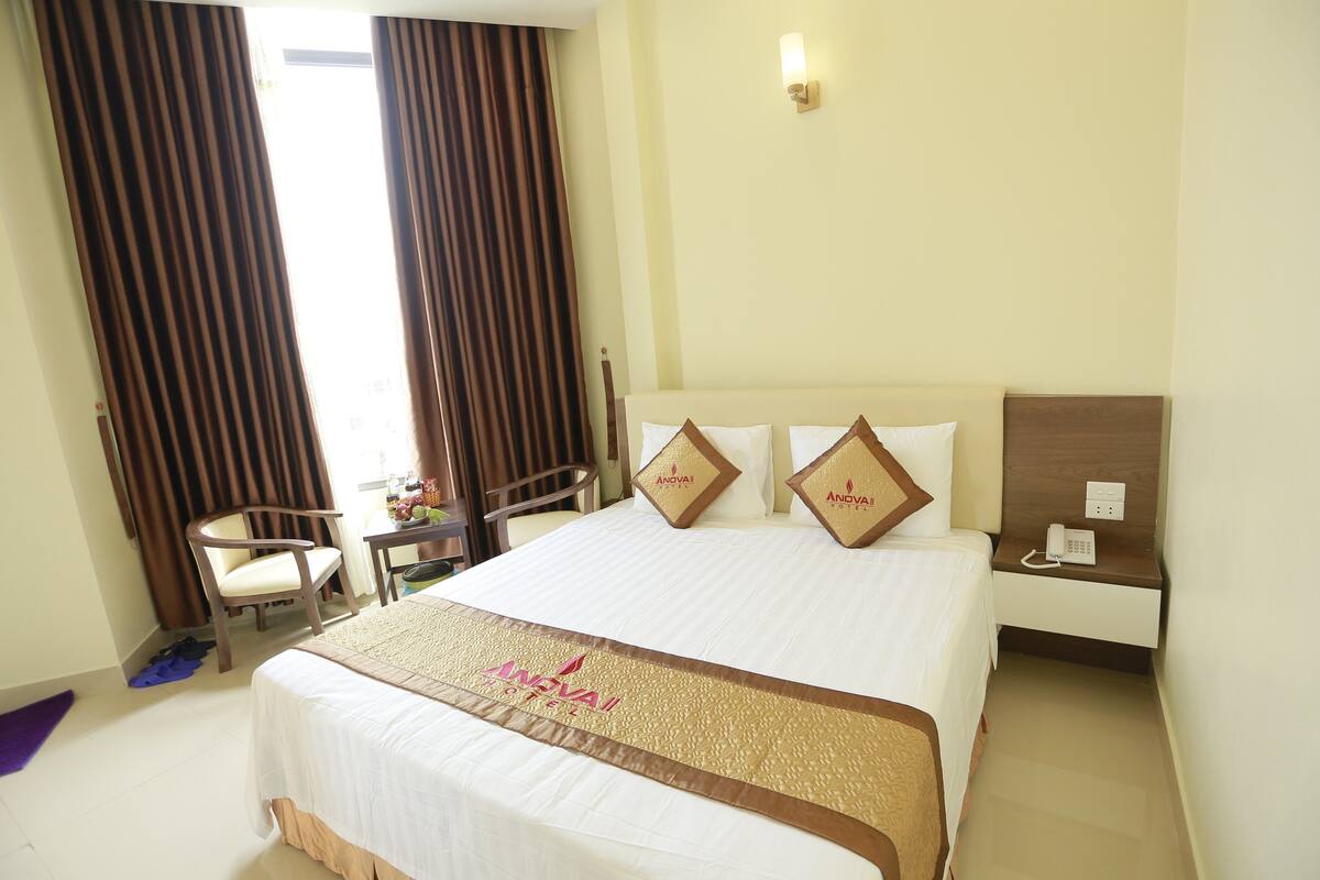 A hotel room with a large bed, two pillows with a logo, a bedside table with a phone, and a chair.
