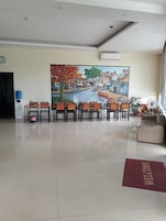 Uma área de recepção com uma grande pintura de uma cena de rua, algumas cadeiras de madeira, um dispensador de água e um tapete de boas-vindas.
