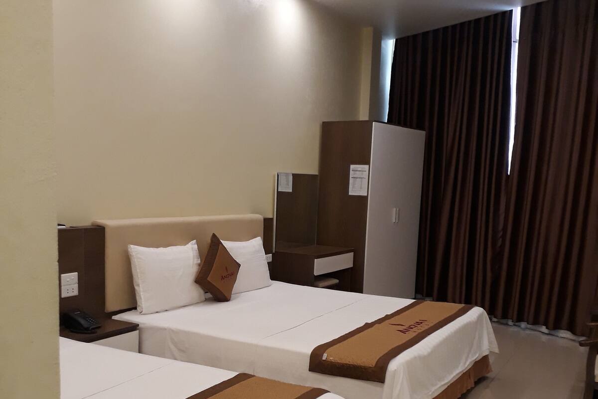 A hotel room with two beds, each with a brown bed runner and white bedding. There is a desk with a chair, a wardrobe, and a telephone on the left side.