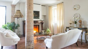 Un salon avec deux fauteuils blancs, une table basse en verre, une cheminée et une grande fenêtre avec des rideaux.