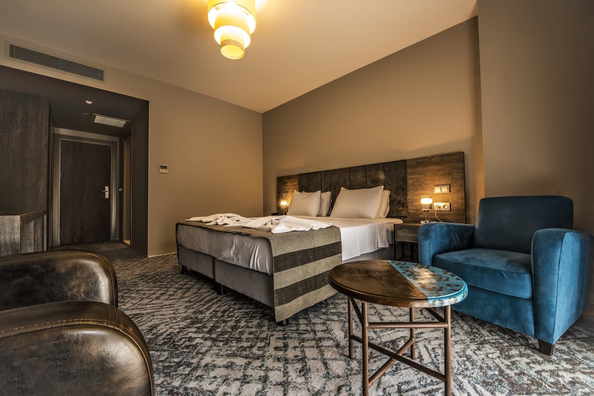 Habitación de hotel con una cama, un sillón azul, una mesita redonda y un cabecero con luces empotradas.