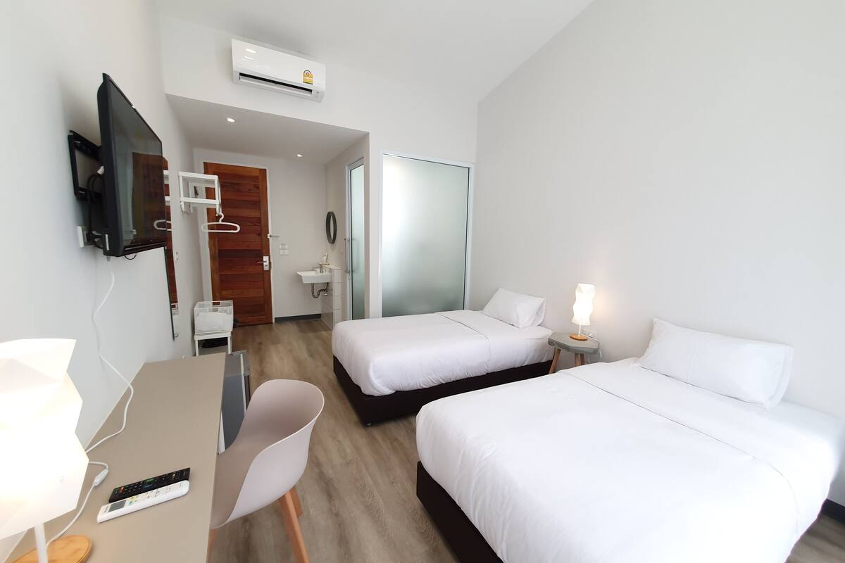 A hotel room with two beds, a desk with a chair, a TV, a bedside table with a lamp, a mirror, and a bathroom visible through an open door.
