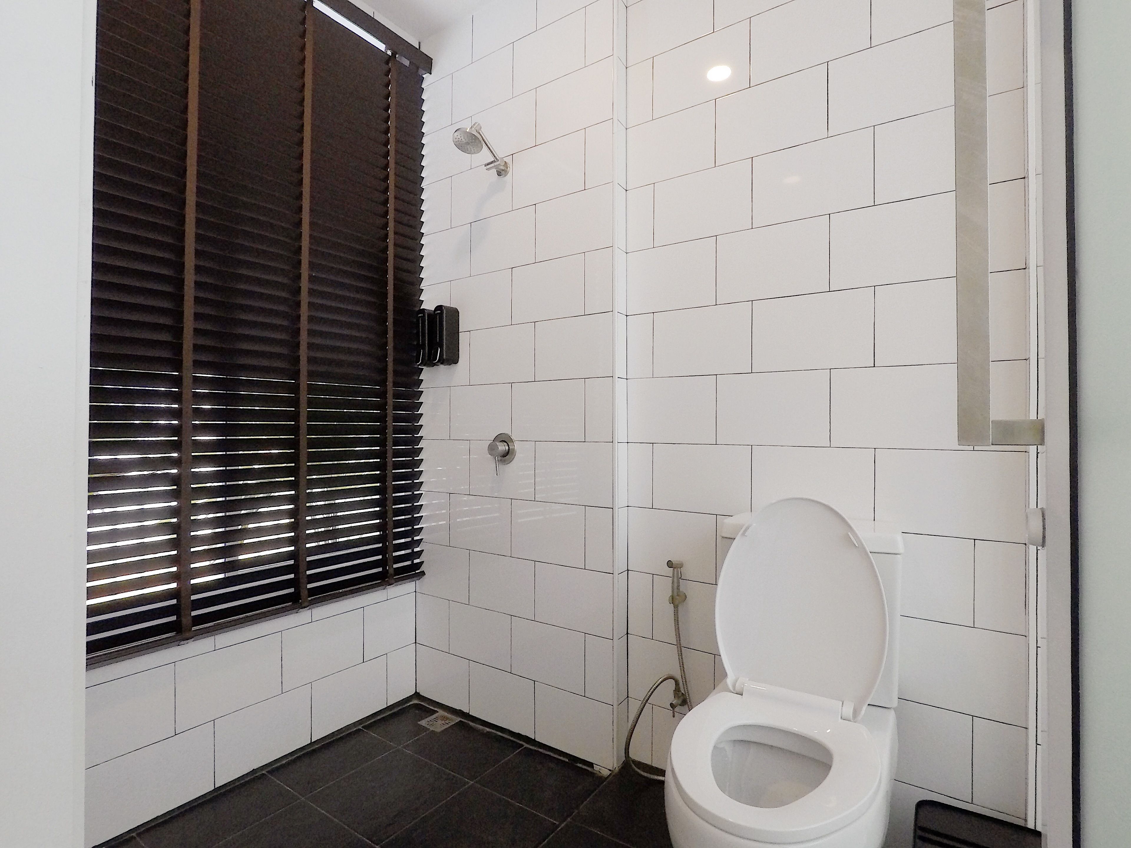 A small, clean bathroom with white tiles, a toilet, a shower, and a window with blinds.