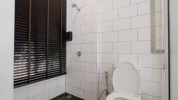 A small, clean bathroom with white tiles, a toilet, a shower, and a window with blinds.