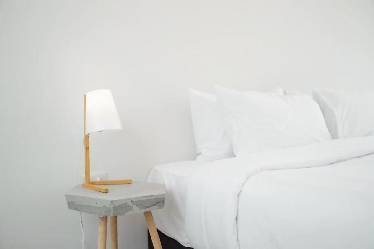 A minimalist bedroom with a bed, white bedding, and a small bedside table with a lamp.