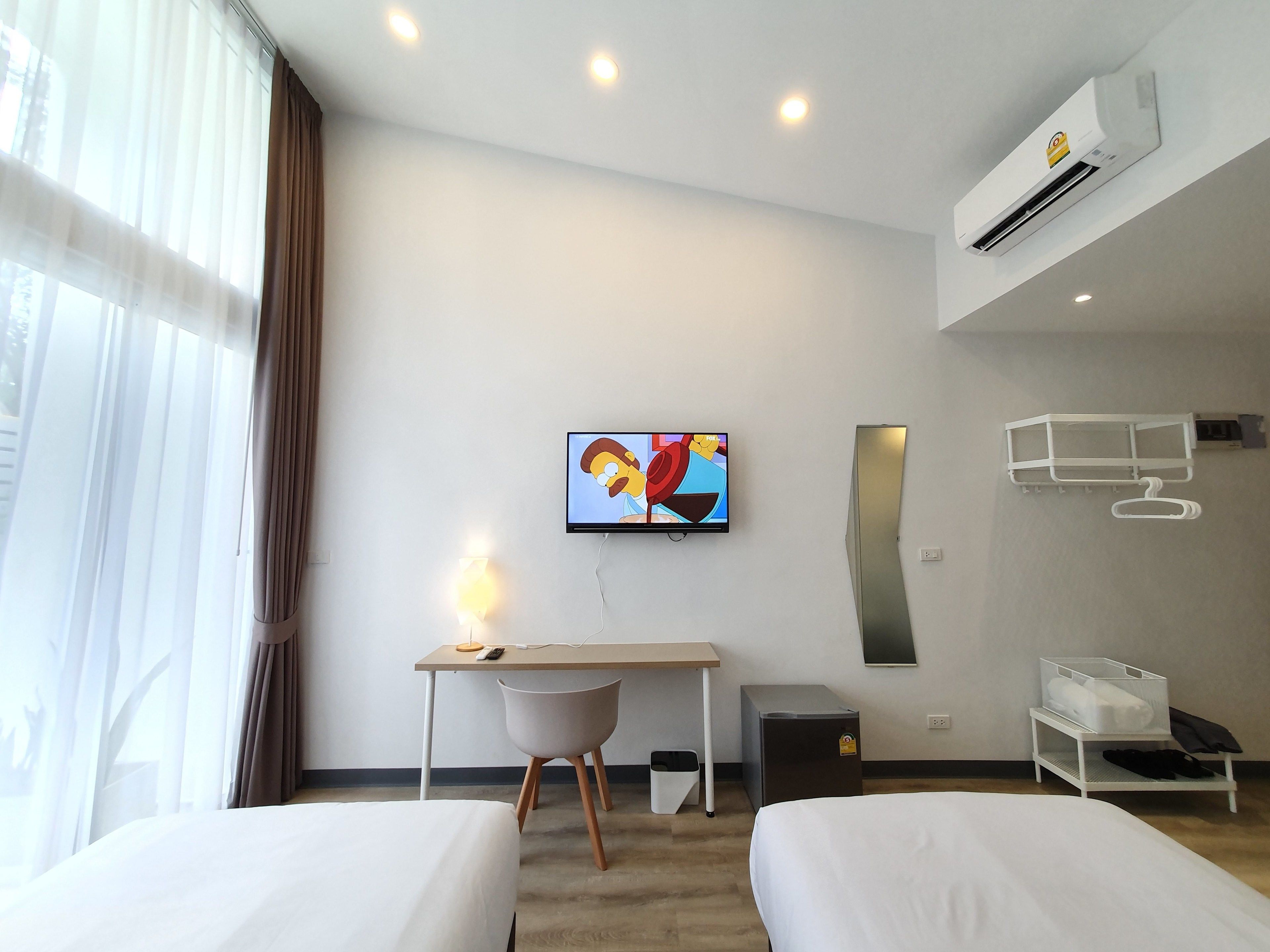 A hotel room with two beds, a desk with a chair, a television, an air conditioning unit, a waste bin, and a storage unit with a hanging rack.